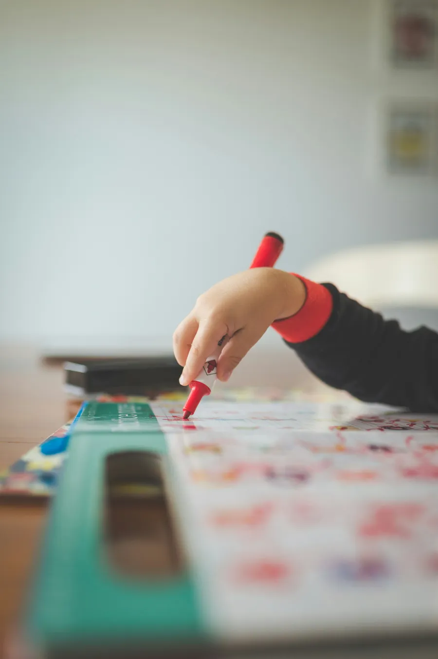 A child's hand holding a red crayon over a colorful worksheet