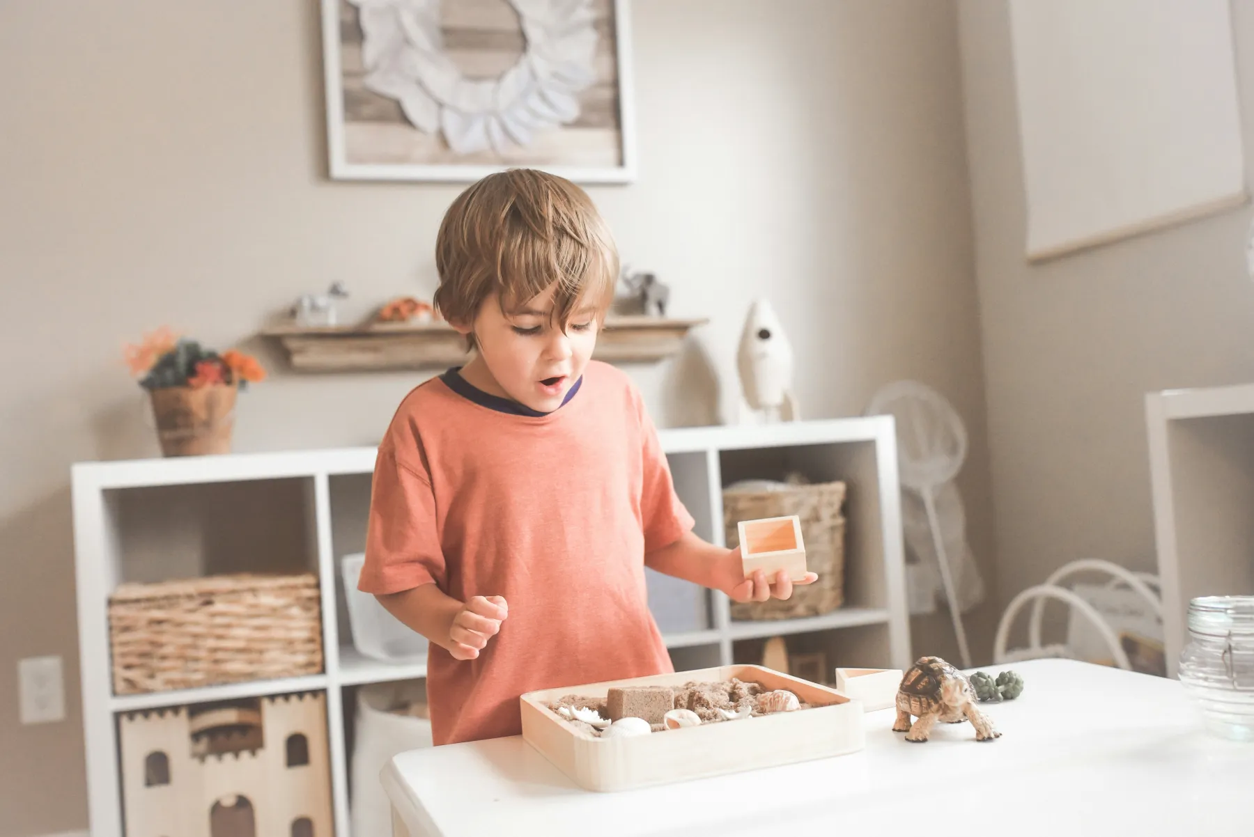 A child exploring a tray of small objects in a bright, cubby-lined learning room